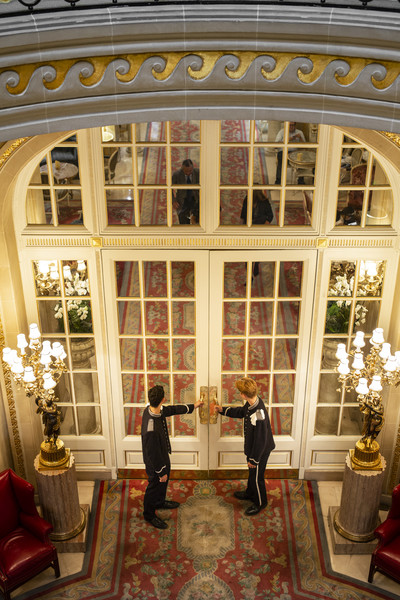 Two doormen in elegant uniforms open the grand entrance doors of The Ritz London, welcoming guests into the opulent surroundings. The scene is framed by gilded detailing, plush red seating, and warm lighting, evoking a sense of timeless luxury.